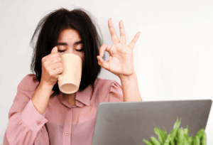 Women with messy black hair sitting at laptop drinking coffee and doing on OK sign with left hand