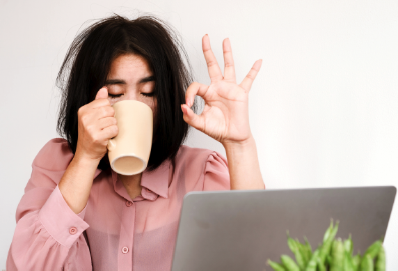 Women with messy black hair sitting at laptop drinking coffee and doing on OK sign with left hand